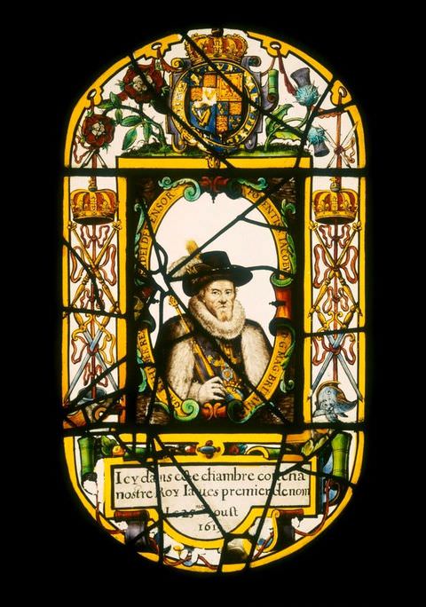 Colour photograph. A photograph of an oval, backlit stained-glass window featuring a colourful, intricate design adorned with plants, flowers, and crowns. Within an oval in the centre of the design is a head to shoulder portrait of a mature man with a beard, wearing a fur-trimmed coat, a tall hat and a stiff white collar.