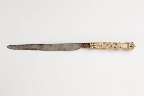Colour photograph. A long, flat knife is photographed against a white background, seen side on. The blade is made from silver coloured metal and contains elements of rust. The handle is carved from a white, ivory-like material, and depicts a standing figure wearing a crown, which has tiny coloured spots on it.
