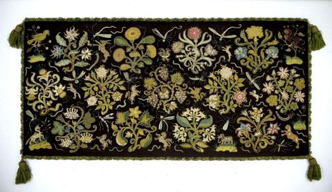 Fabric cushion. A document of a black patterned cushion cover against a white background. The cushion cover is filled with green, pink and white plant and flower motifs, has a green, frilled trim around its edge, and each corner features a green tassel.