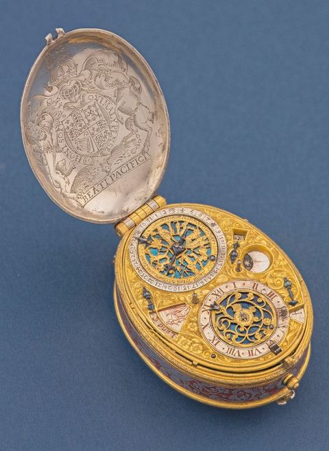 Colour photograph. An oval object with a lid on top is photographed against a blue background. The lid has been lifted to reveal the inside of the object. The inner, upper lid contains the royal British insignia, while the object has circular, mechanical, and numerical details resembling a clock or compass.
