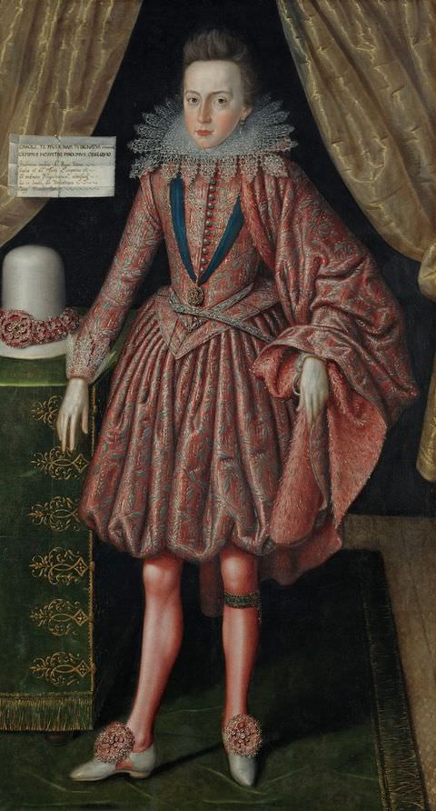 Painting. A full-length portrait of a young man dressed in traditional clothing with a corseted waist and a full, knee-length skirt, topped with an indulgent cape draped over one arm. He wears red tights and bejewelled white shoes. An interior behind him is decorated with green velvet and gold satin, while a tall white hat sits on a table to the left.
