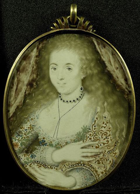 Painting. An oval miniature painting of a woman, showing her head, neck, and decolletage, with arms folded out in front, contained within a gold frame. She wears a pale dress with intricate, floral embroidery and a low, open neckline, a high, choker necklace, and has loose strands of wavy blonde hair flowing down her shoulders.