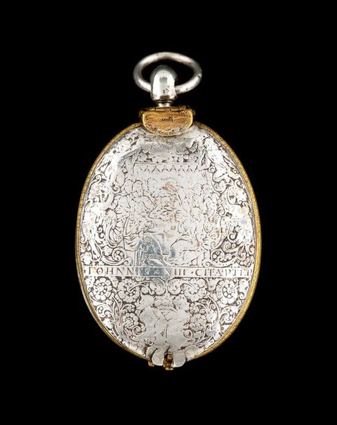 Colour photograph. A document of an oval, silver miniature with the lid closed shut, and a small, circular loop on the top. The cover is decorated with intricate, ornate areas of pattern and a strip of text running from left to right just below the centre.