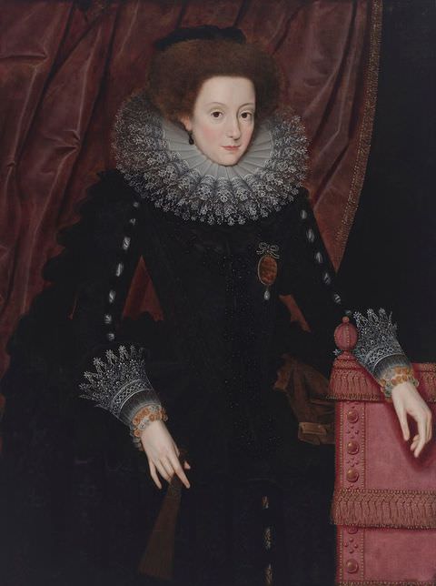 Painting. A portrait of a woman seen to the waist, wearing a black dress with white lace cuffs, and a stiff, white lace collar. She wears a large red brooch on her right chest area. Behind her is a dark red curtain, slightly swept to one side. Her left arm rests on a pale red piece of furniture at waist height. 