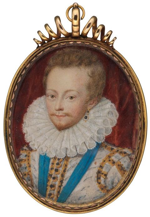 Painting. An oval, miniature painting with a gold frame, depicting a portrait of a man seen from head to neck. He is wearing a fitted white jacket with gold details, a blue sash, and a white neck ruff. Behind him is a dark red backdrop.