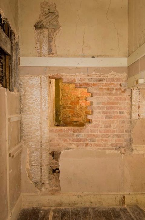 Colour photograph. A section of a room is documented, showing worn out wood floorboards, sections of bare, red brick where plasterboard has worn away, and missing sections of wall with bare wooden timbers visible beneath.  