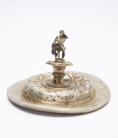 Colour photograph. This photograph documents a golden, decorative object against a white backdrop. The object is circular in design and features embossed, organic patterns. A shape resembling a candle holder emerges from the object’s centre, with a small figure stand on its top.