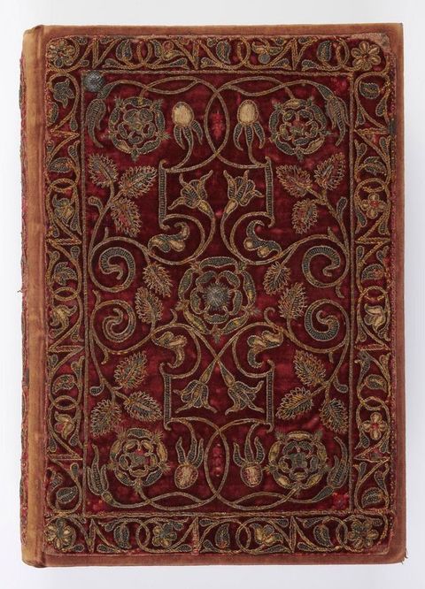 Embellished book cover. Late sixteenth-century Bible with a deep red velvet binding densely embroidered in gold and silver thread, featuring symmetrical scrolling vines, flowers, and Tudor rose motifs within a decorative border; the textile cover shows wear along the edges, indicating age and use.