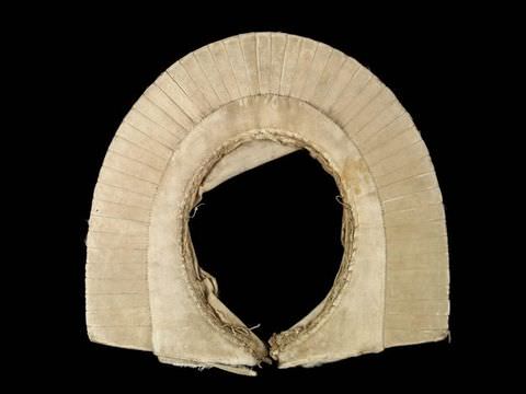 Colour photograph. An old-fashioned, white, pleated garment resembling a collar is photographed laid out flat against a black backdrop. Loose, rough stitches are visible in the inner curves of the item.