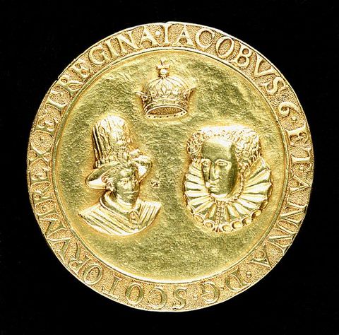 Colour photograph. A circular golden coin documented against a black backdrop. The coin features two heads facing us – a man and a woman – with a crown suspended in the space above them. A passage of text encircles the circumference of the coin.