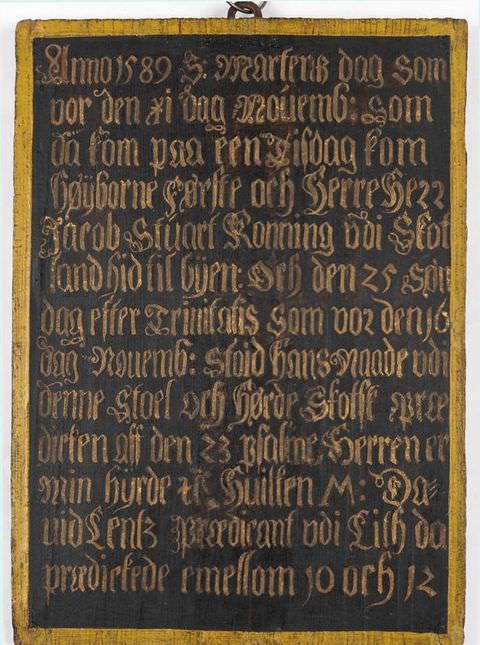 Colour photograph. Documentation of a black plaque resembling a chalk board inscribed with gold, calligraphic lettering, photographed against a white background. The plaque is surrounded by a simple gold frame and has a small hoop at the top.